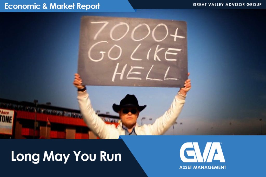 Person wearing a cowboy hat and sunglasses holds up a chalkboard sign reading '7000+ GO LIKE HELL' at a stadium backdrop, with an 'Economic & Market Report' banner at the top.